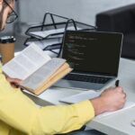 Człowiek z brodą studiuje z książką w pobliżu laptopa z komputerowym kodowaniem na ekranie w nowoczesnym biurze