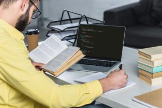 Człowiek z brodą studiuje z książką w pobliżu laptopa z komputerowym kodowaniem na ekranie w nowoczesnym biurze