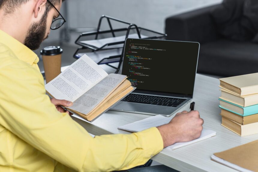 Człowiek z brodą studiuje z książką w pobliżu laptopa z komputerowym kodowaniem na ekranie w nowoczesnym biurze
